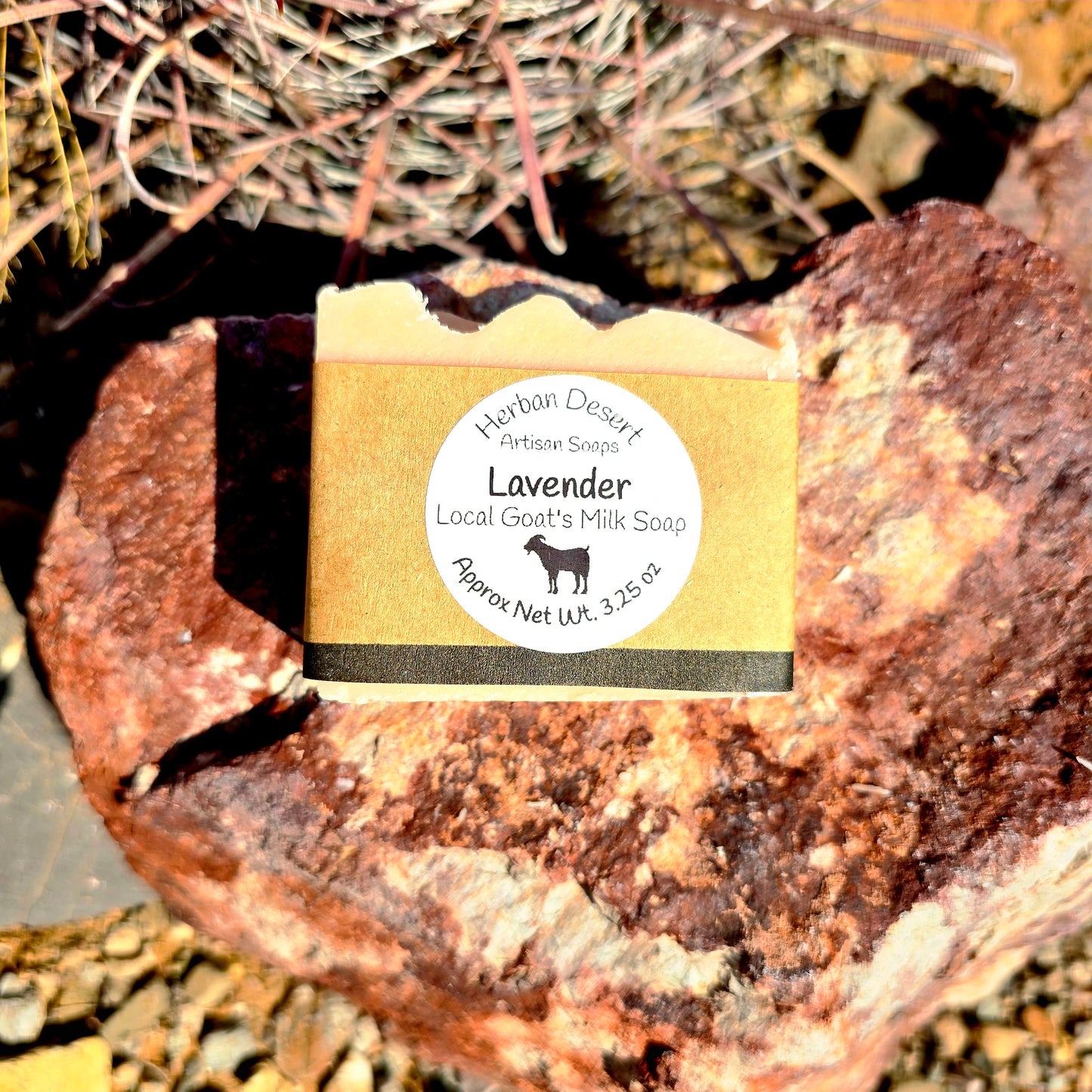 A bar of goat milk natural soap with lavender essential oil, labeled 'Herban Desert', placed on a natural rock surface.
